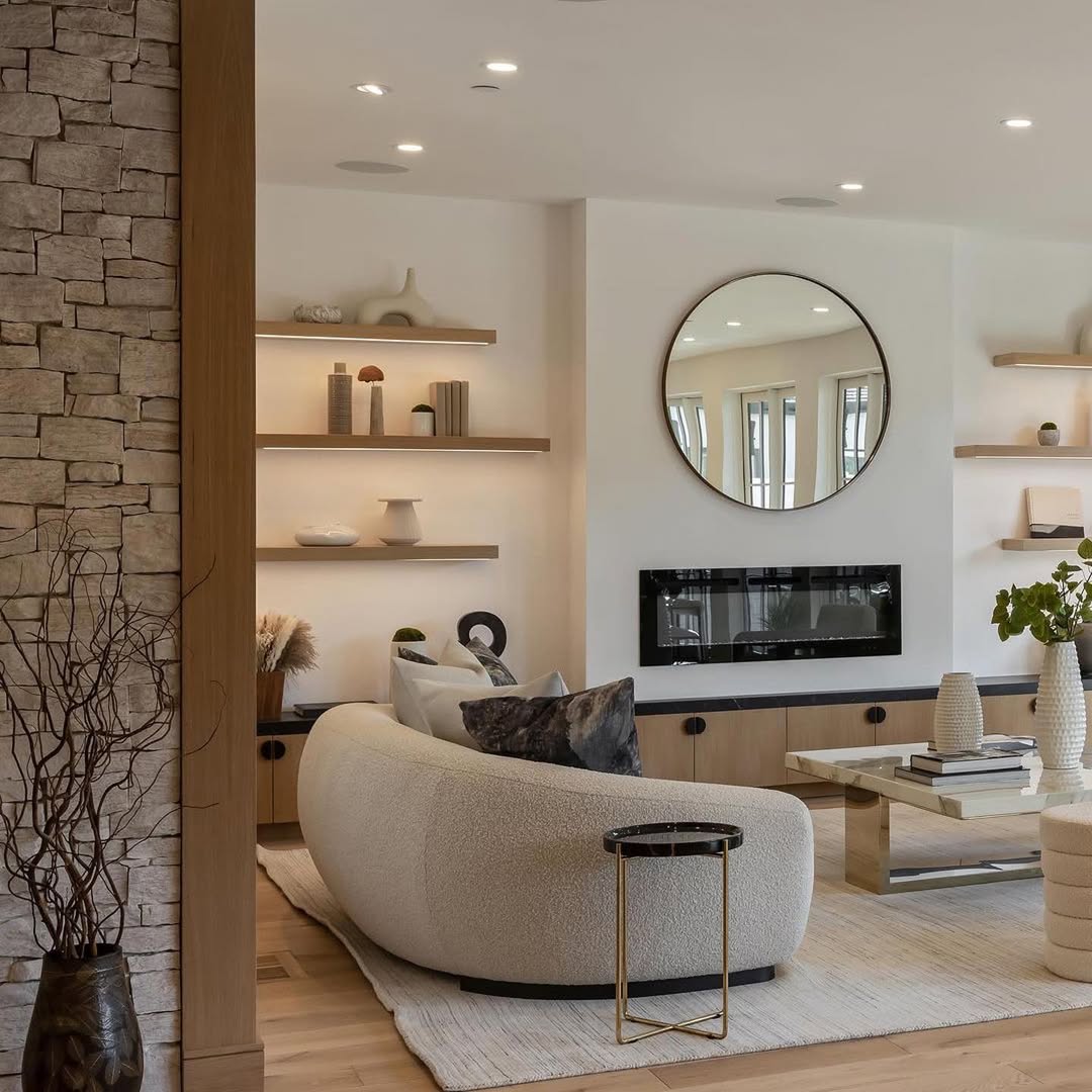 Modern living room ideas come to life with a curved sofa, round mirror above a black fireplace, floating shelves with decor, and a neutral palette. Stone accent wall and wooden flooring complete this organic modern living room design.