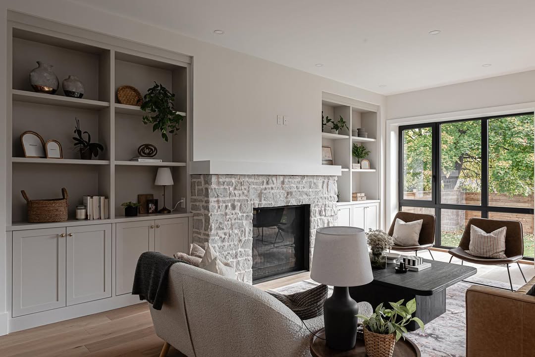 Modern living room with a stone fireplace and stylish fireplace built-ins, complemented by neutral-toned furniture. A large window fills the space with natural light and frames views of trees outside—great inspiration for fireplace ideas.