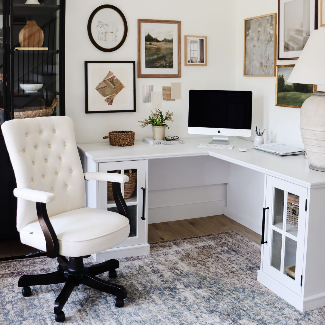 A home office features a sleek white L-shaped computer desk with an iMac, an upholstered swivel chair, framed art on the wall, and decorative items—perfect for inspiring new desk ideas.