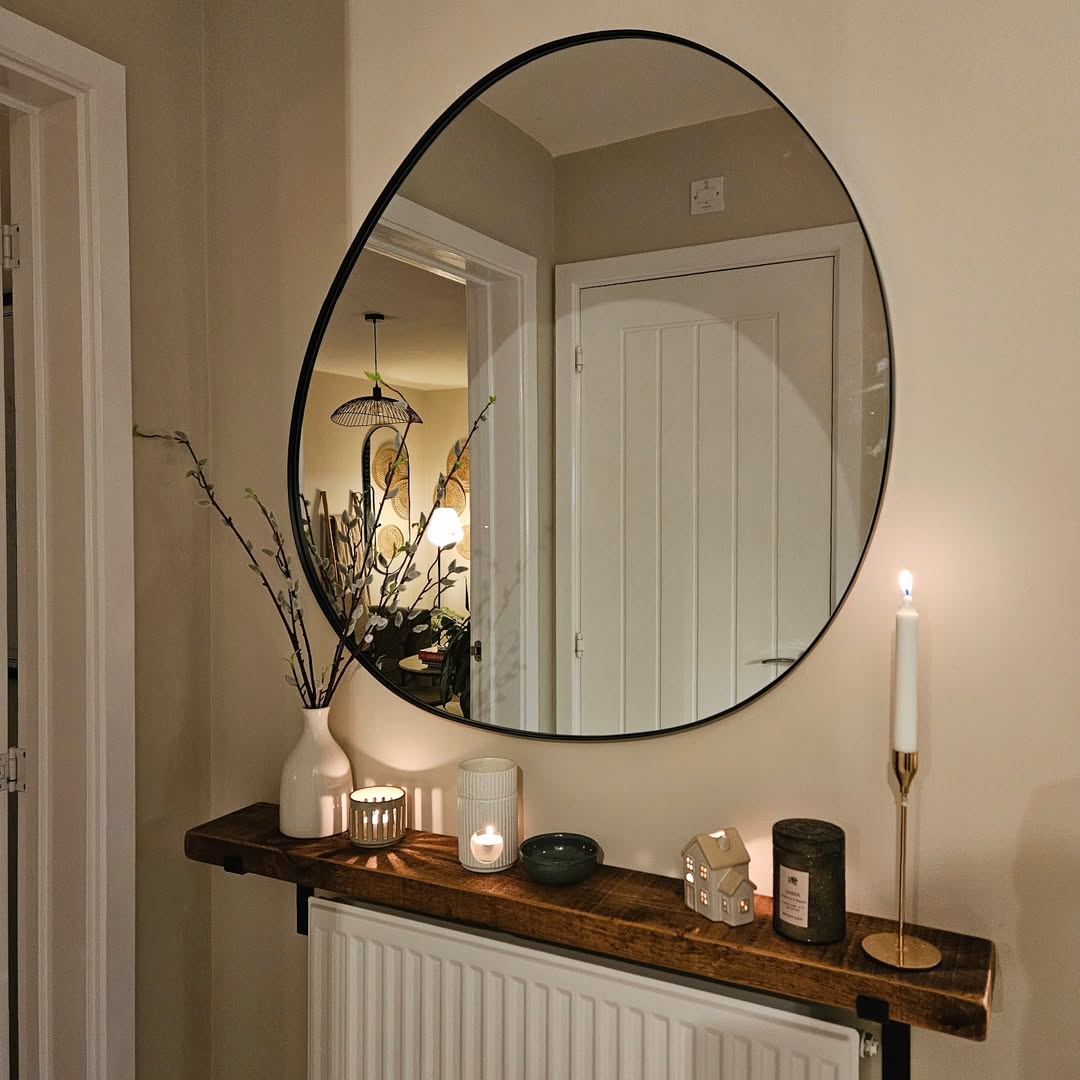 A round mirror hangs above a wooden shelf, offering console table decor inspiration with a vase of branches, candles, a small house ornament, and a lit candleholder; doorway reflections are visible.