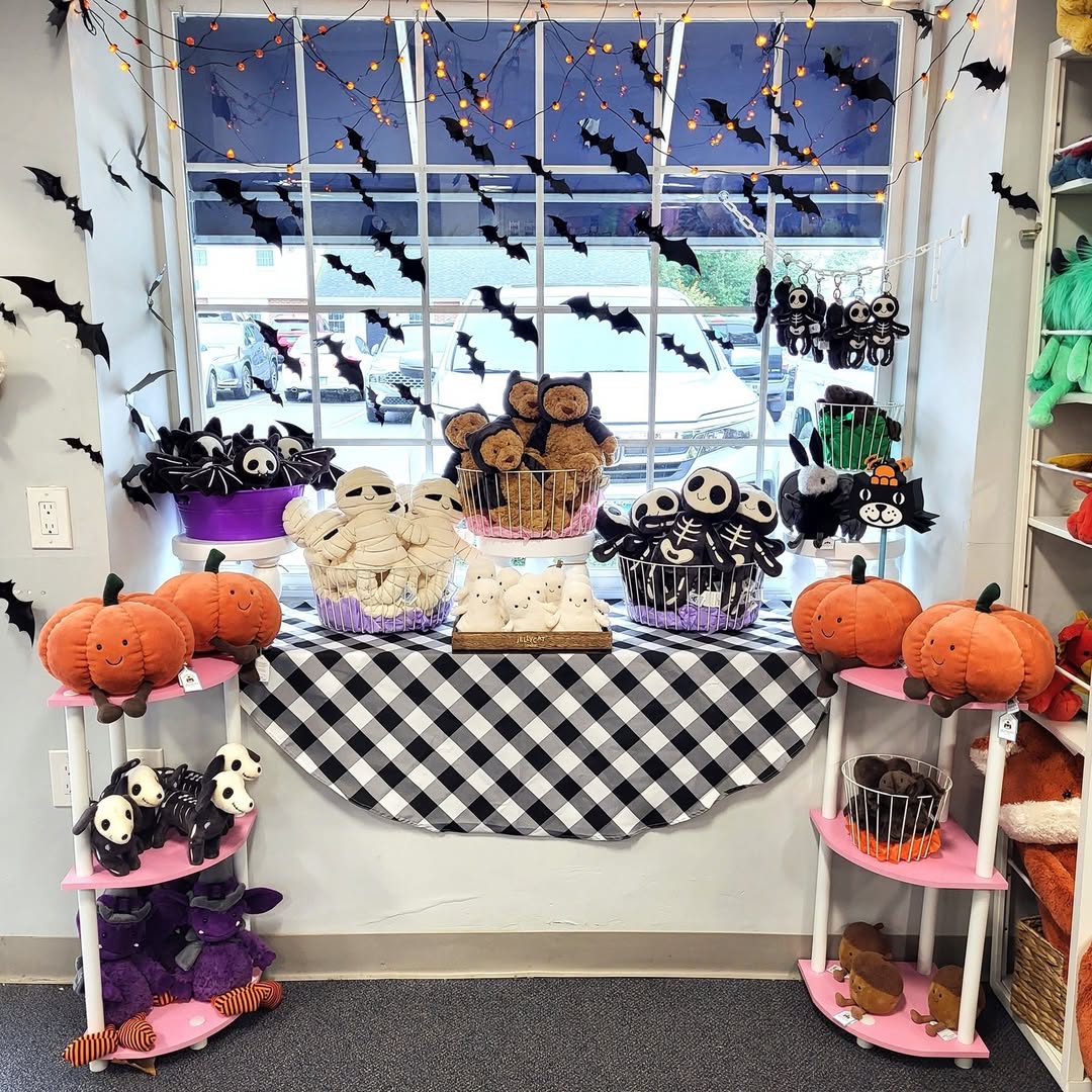 A store window display features Halloween plush toys—pumpkins, mummies, cats, skeletons, and bats—arranged on shelves and a table with a black-and-white checkered cloth, offering creative Halloween window decoration ideas.