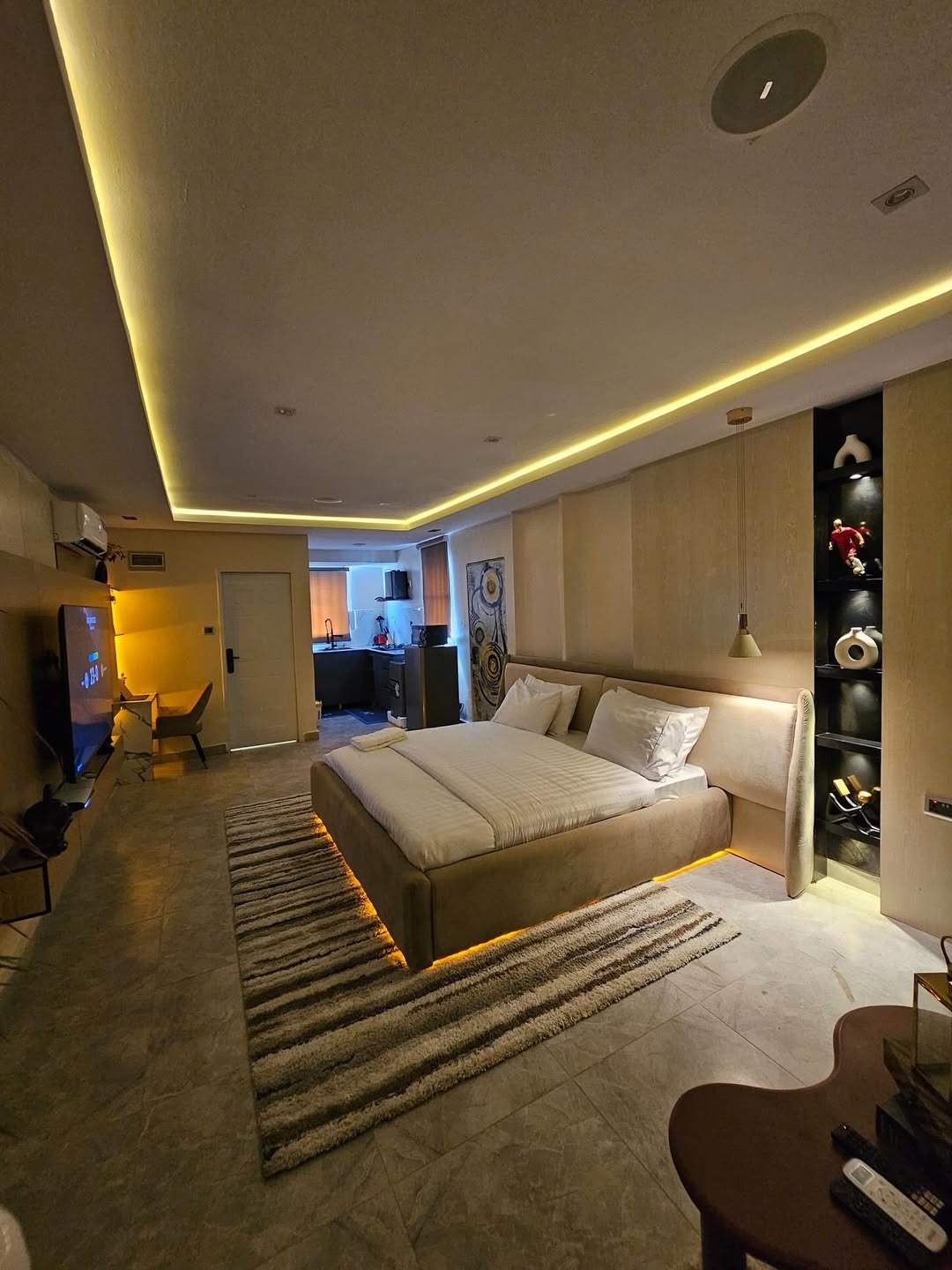 Modern bedroom with ambient lighting, a large bed, wall-mounted TV, open shelving, and a connected seating area with a desk and yellow chair—perfect studio decor inspiration for smart small space design.