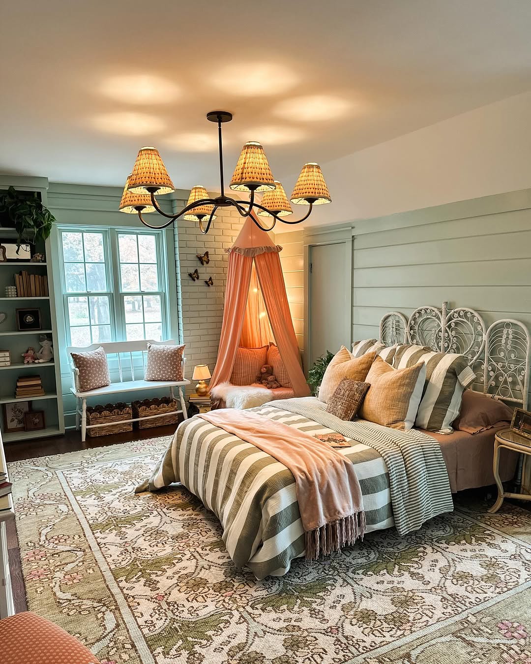 A cozy sage green farmhouse bedroom with a striped bed, canopy, chandelier, area rug, green built-in shelves, and a window seat with pillows.