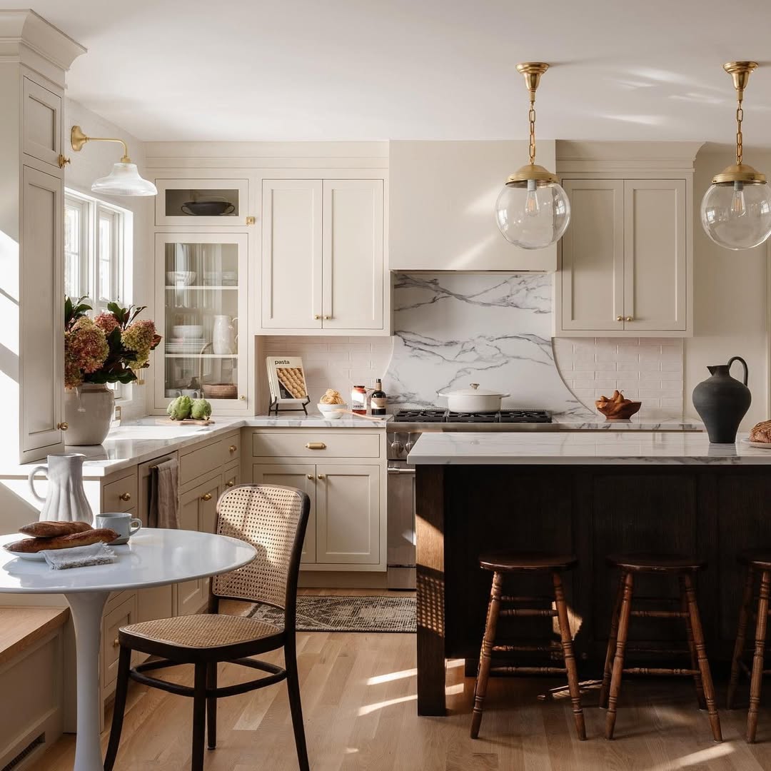 Bright kitchen with white cabinets, marble backsplash, and a dark island with wooden stools—this space reflects stunning kitchens and kitchen color ideas that are sure to inspire kitchen trends 2025. Decor includes vases, flowers, and pendant lights with natural light.