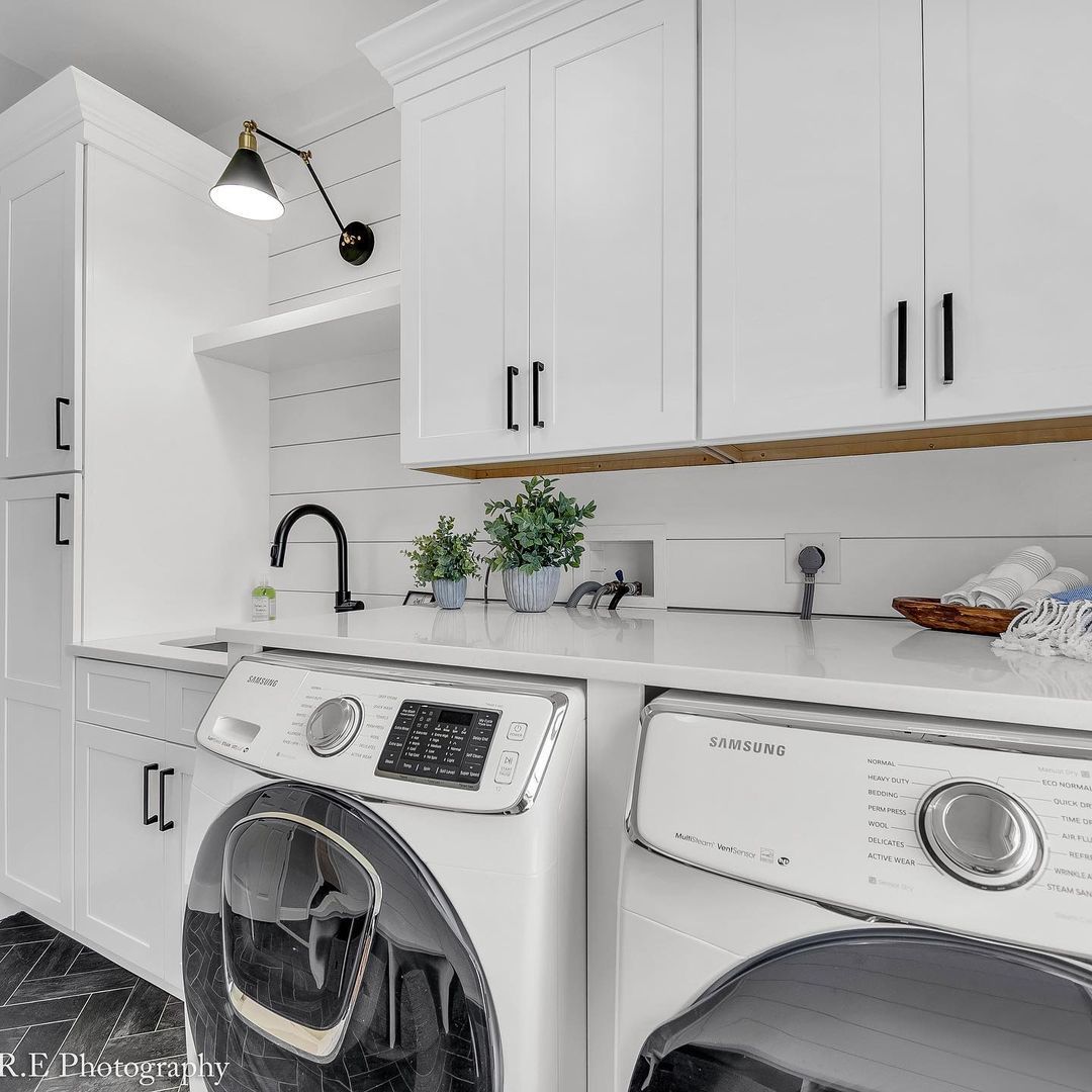 Modern laundry room with white laundry cabinets, black hardware, washer and dryer, countertop, wall light, and decorative plants on a white backsplash—perfect inspiration for stylish laundry room cabinet ideas.