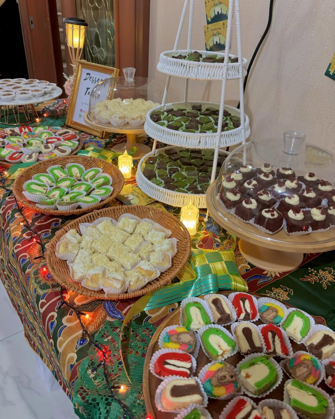 A dessert table with assorted traditional pastries and party desserts arranged on trays and stands, decorated with fairy lights and patterned cloth—perfect for inspiring new dessert table ideas.