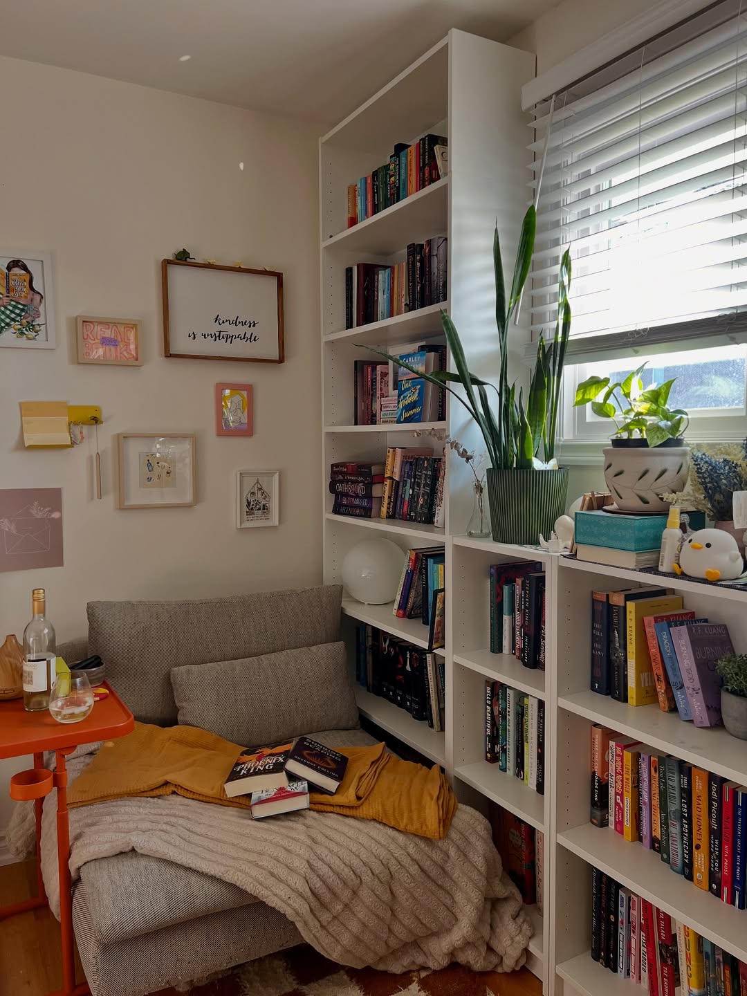 A cozy reading nook with a gray chair, blanket, and books, surrounded by white bookshelves filled with books and plants, offers inspiring Book Nook Ideas next to a window with blinds.