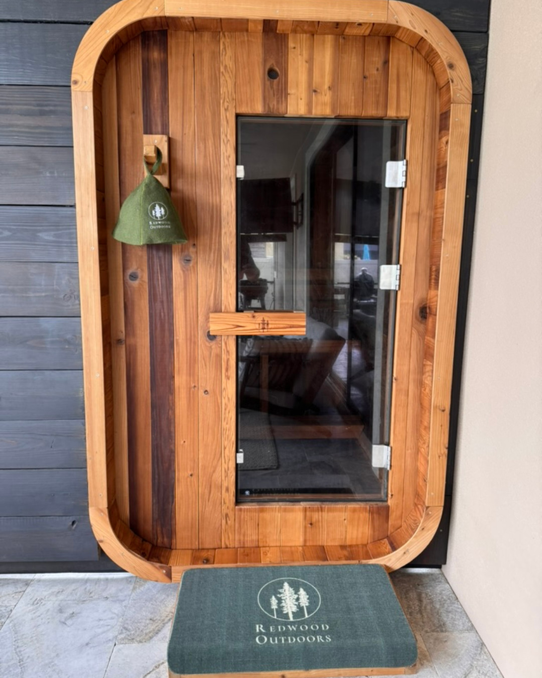 A wooden outdoor sauna with a glass door, a green cap hanging on a hook, and a green