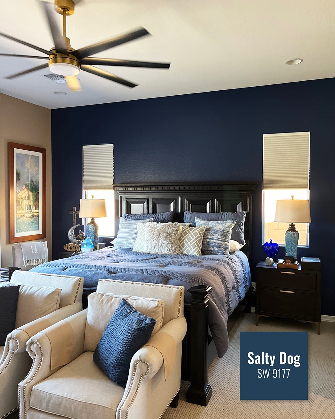 A modern bedroom for Interior Lovers featuring a dark wood bed, blue and beige bedding, two beige chairs, matching nightstands, lamps, and a navy accent wall labeled
