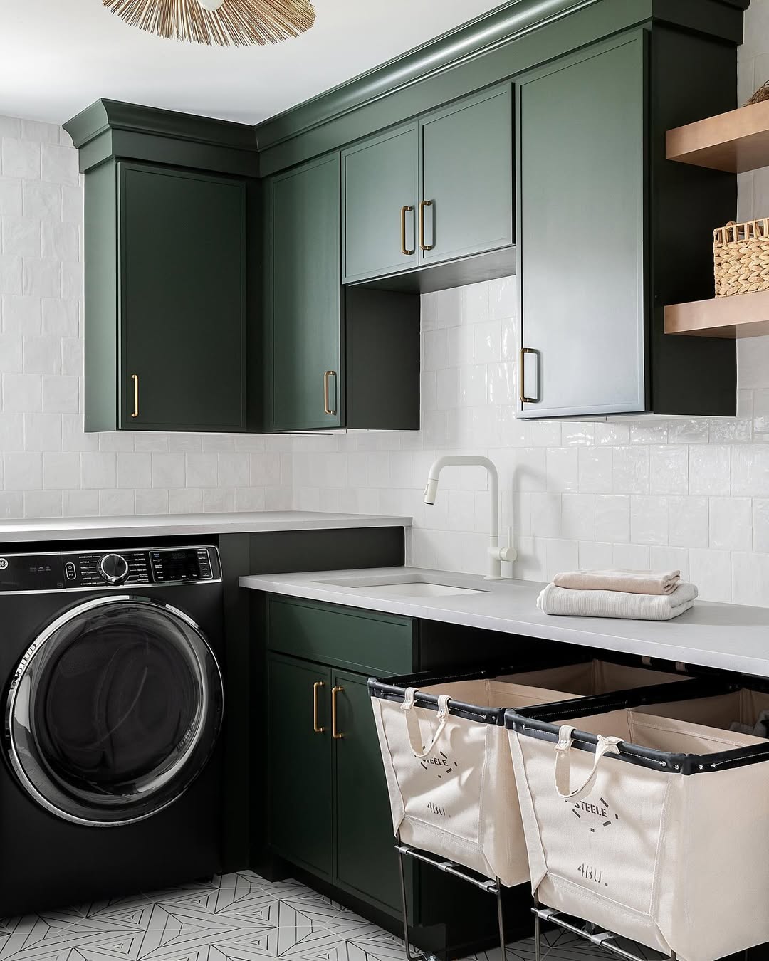 Modern laundry room with dark green cabinets, a front-loading washer, white countertops, subway tile backsplash, open wooden shelves, and two canvas laundry bins—perfect inspiration for stylish Laundry Room Cabinet Ideas.