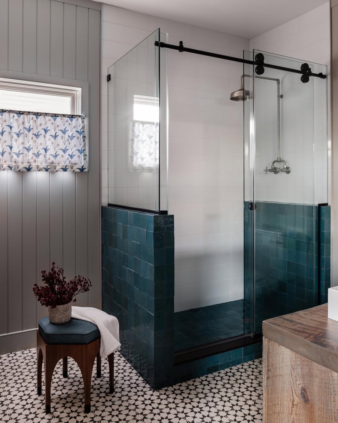 A bathroom with a glass-enclosed shower featuring blue-green tiles, patterned floor tiles, a wooden stool with a vase, and a window with a floral curtain—a standout shower design for fresh bathroom ideas.