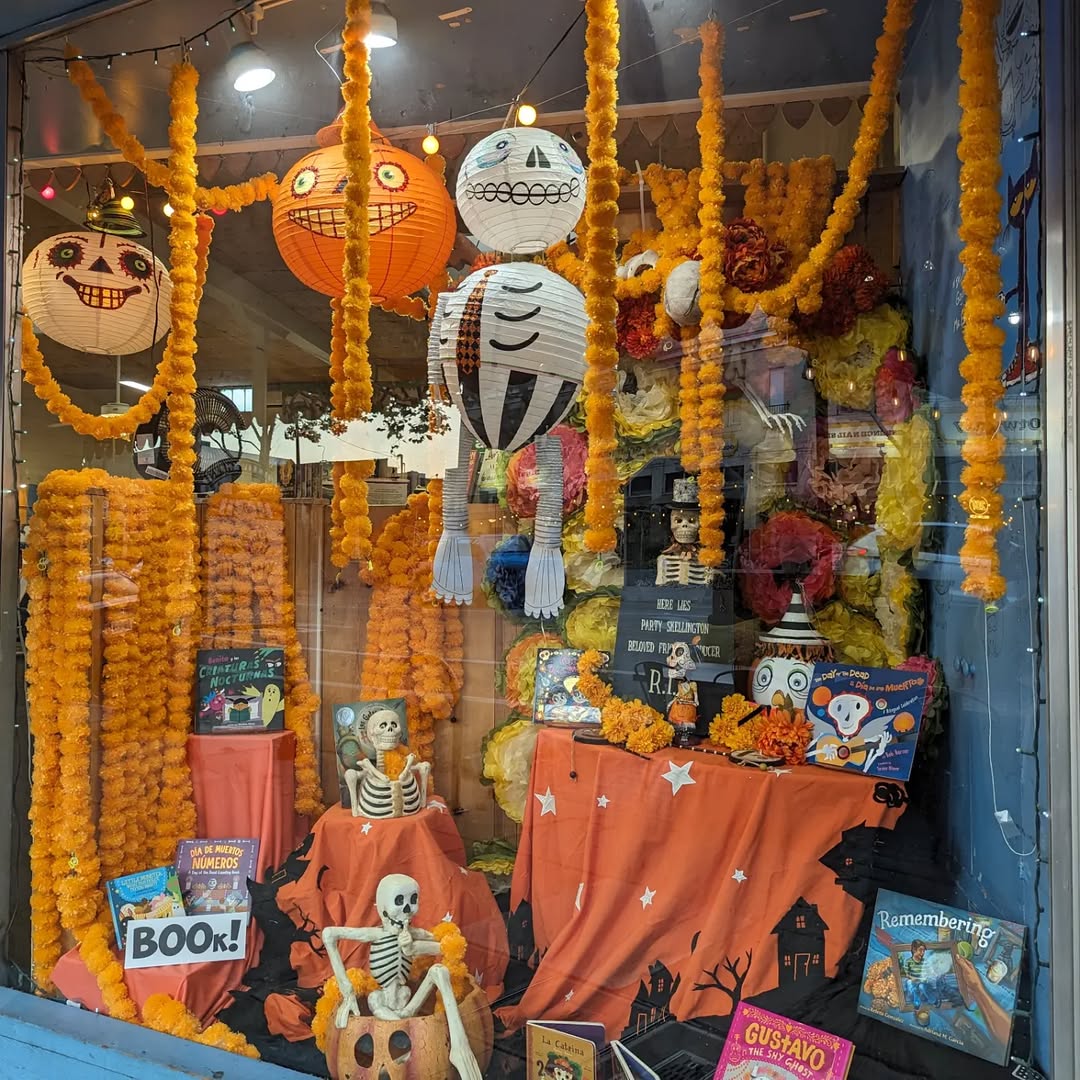 A store window display decorated for Día de los Muertos with marigold garlands, skeleton figures, papel picado, colorful lanterns, children