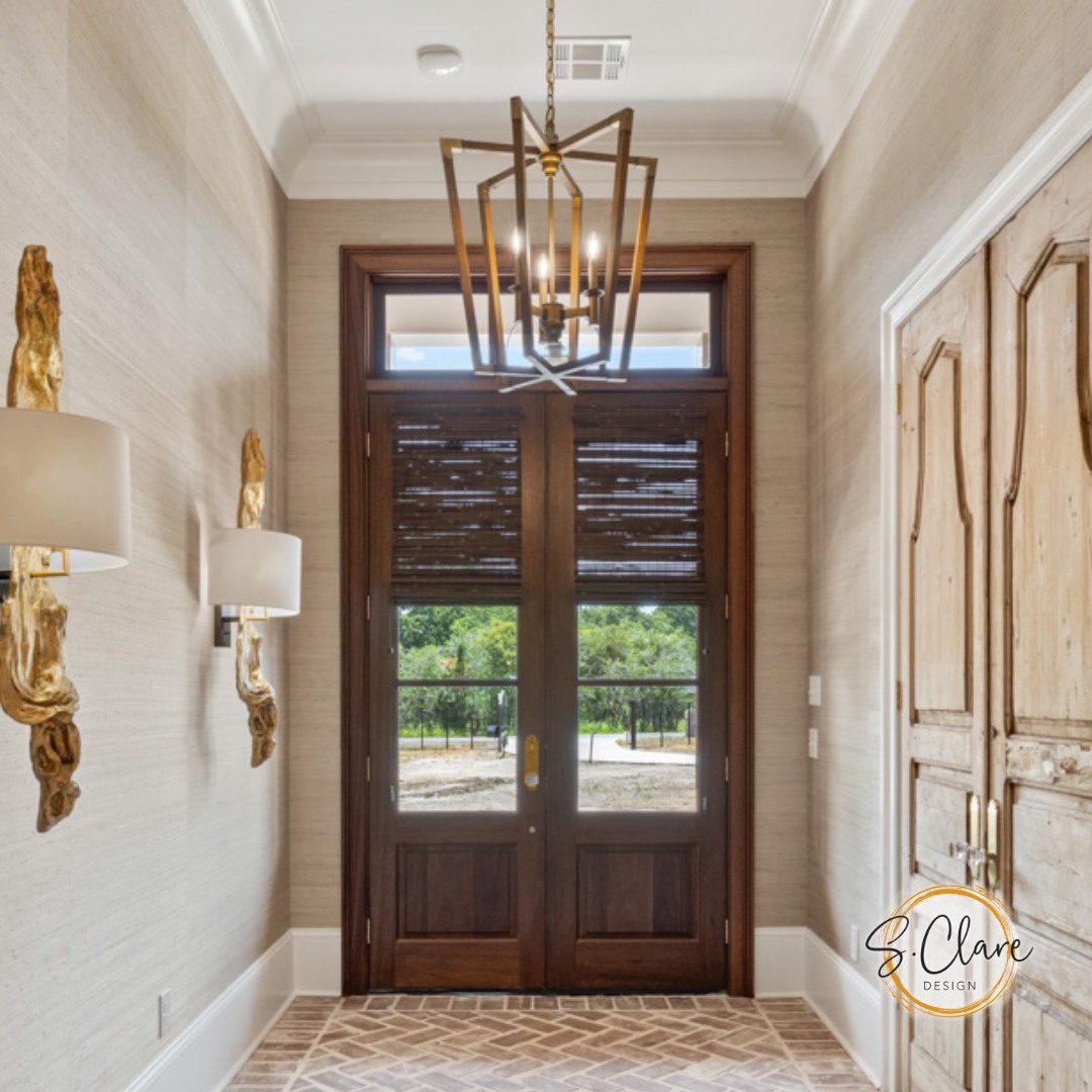 A hallway with a stunning double door entryway, a geometric chandelier, wall sconces, textured wallpaper, and a view of greenery outside—perfect inspiration for elegant entryway ideas.
