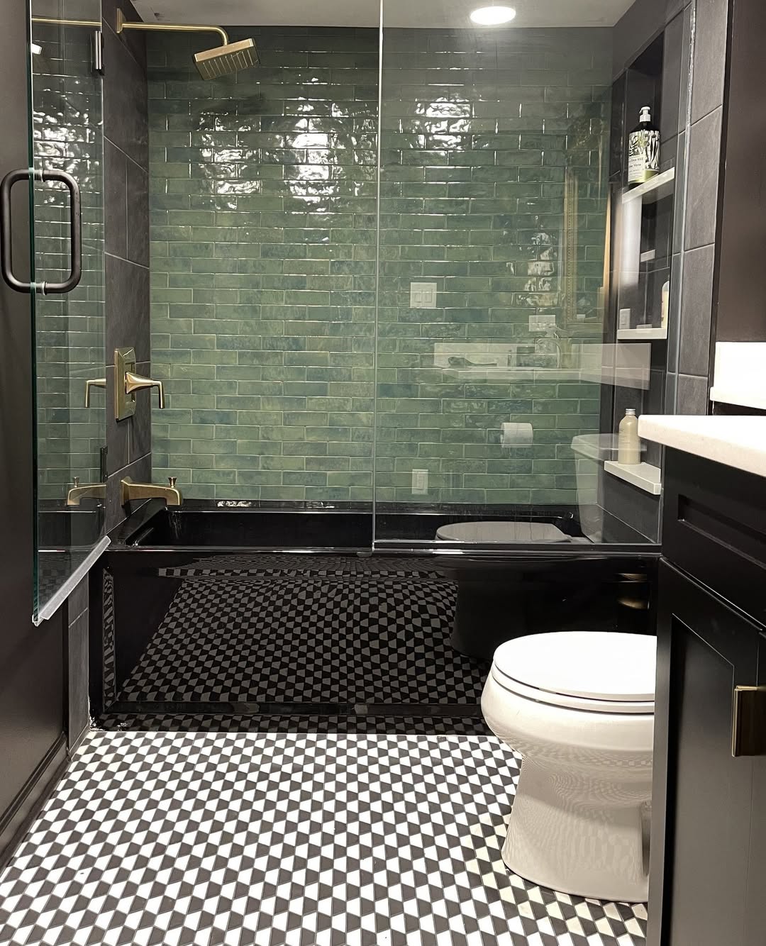 Modern spa bathroom with a glass shower, green subway tile wall, brass fixtures, black-and-white geometric floor tiles, toilet, and built-in shelving—perfect for fresh bathroom design inspiration.