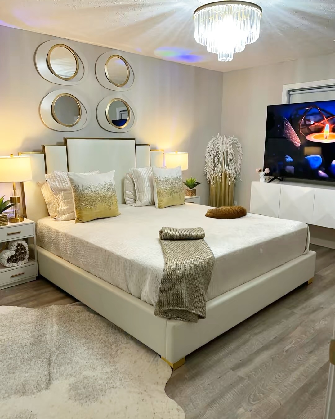 Modern bedroom with a large white bed and stylish bedroom headboard, decorative pillows, a folded blanket, round mirrors on the wall, side tables with lamps, and a TV showing a candle flame.