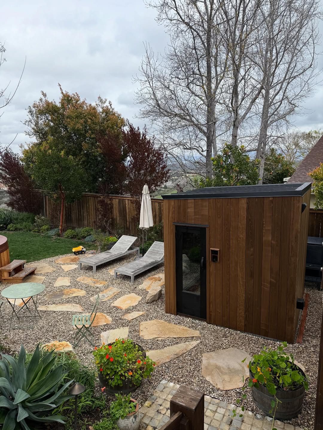 A backyard with a modern wooden shed, two lounge chairs, a side table, large stone pavers, potted plants, and trees along a wooden fence under a cloudy sky—perfect for relaxing or exploring outdoor sauna ideas.