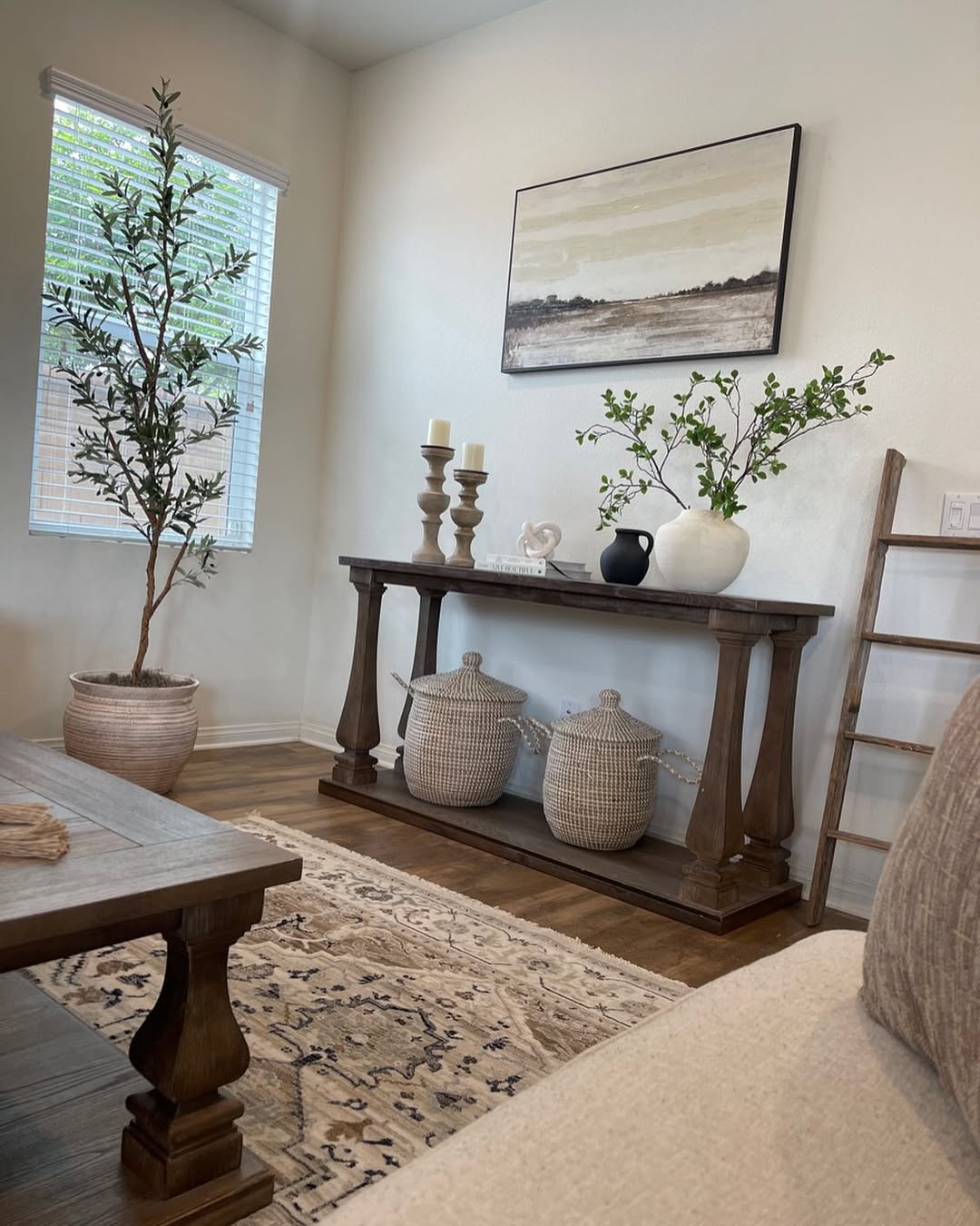 A living room corner with a wooden console table, stylish vase decor, woven baskets, a potted plant, a ladder, and a landscape painting on the wall.