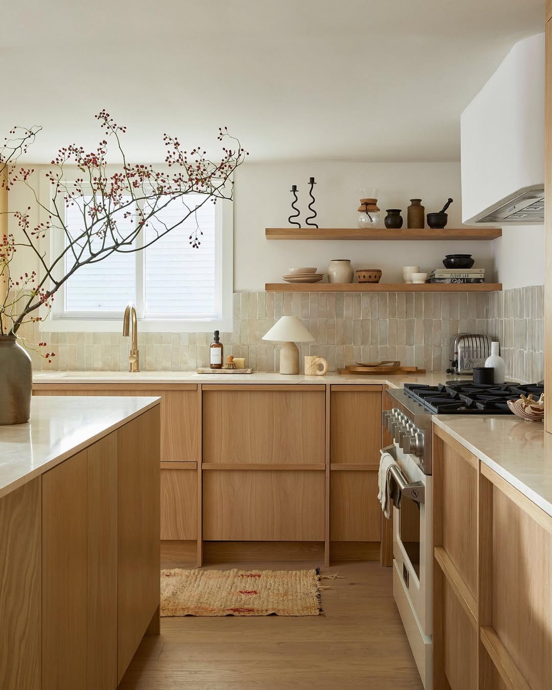Modern oak kitchen with light wood cabinets, open shelves displaying ceramics, a neutral tile backsplash, a large branch in a vase, and a sunlit window above the sink—perfect for inspiring fresh kitchen design ideas.