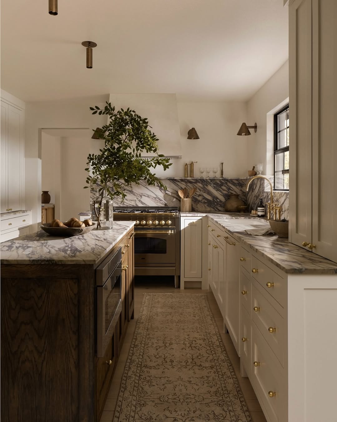 A modern kitchen with white cabinets and marble countertops showcases stunning kitchen colors, gold hardware, a central island, a patterned rug, and a potted branch as decor. Natural light enters through a window.