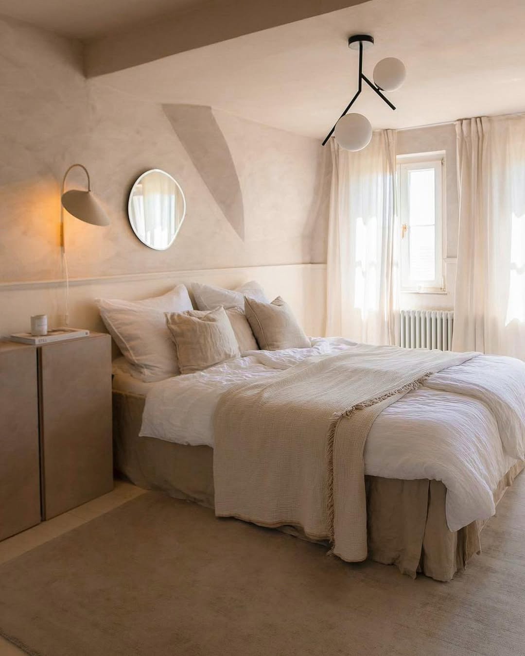 Minimalist bedroom with a smart small bedroom layout, featuring neutral tones, a made bed with white and beige bedding, round mirror, wall lamp, modern ceiling light, compact cabinet, and sheer curtains over the window.