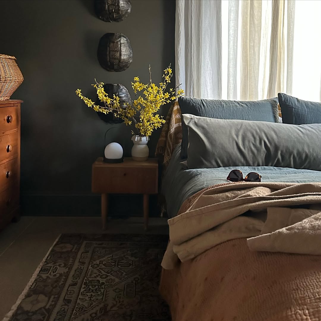 A neatly made bed with gray and beige bedding showcases trendy bedroom color schemes for 2025, beside a wooden nightstand holding yellow flowers, a lamp, and a decorative rug. Light filters through sheer curtains—perfect for interior lovers.