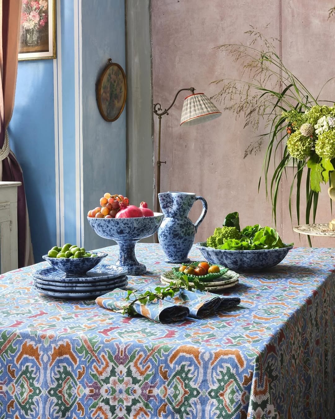 A table set with patterned blue and white dishes holding fruits, vegetables, and a pitcher sits on a colorful tablecloth amid vintage decor and plants, offering inspiring table setting ideas for your home.