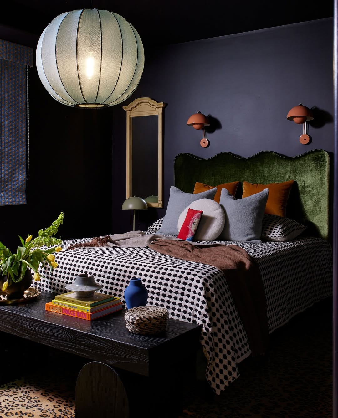 A dark feminine bedroom with moody walls, a green velvet headboard, polka dot bedding, assorted pillows, a wooden bench stacked with books, and a large round paper lantern ceiling light.