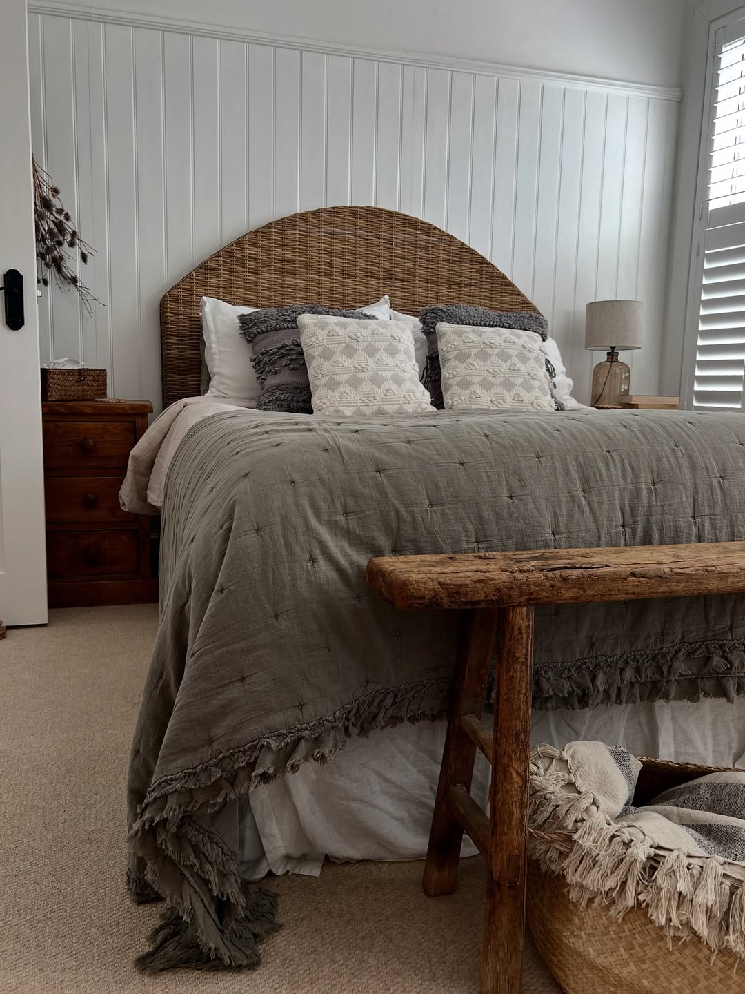 A neatly made bed with a wicker headboard, decorative pillows, and a gray blanket is centered in a cozy, neutral-toned rustic bedroom with wood furniture and white paneled walls, perfect for inspiring your bedroom decor.