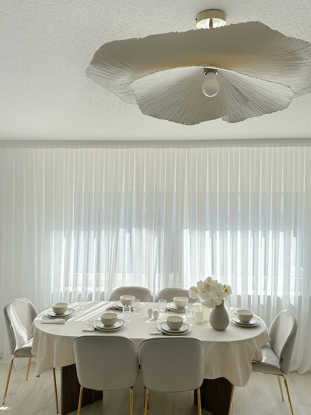 A dining table set for six with white dishes and a vase of flowers showcases elegant table setting ideas beneath a large, textured ceiling light fixture in a bright room with sheer curtains.