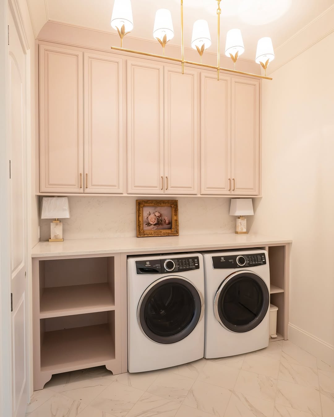 A laundry room with front-loading washer and dryer, beige laundry room cabinets, two lamps, a framed picture, and marble tile flooring—perfect for those seeking fresh laundry room ideas.