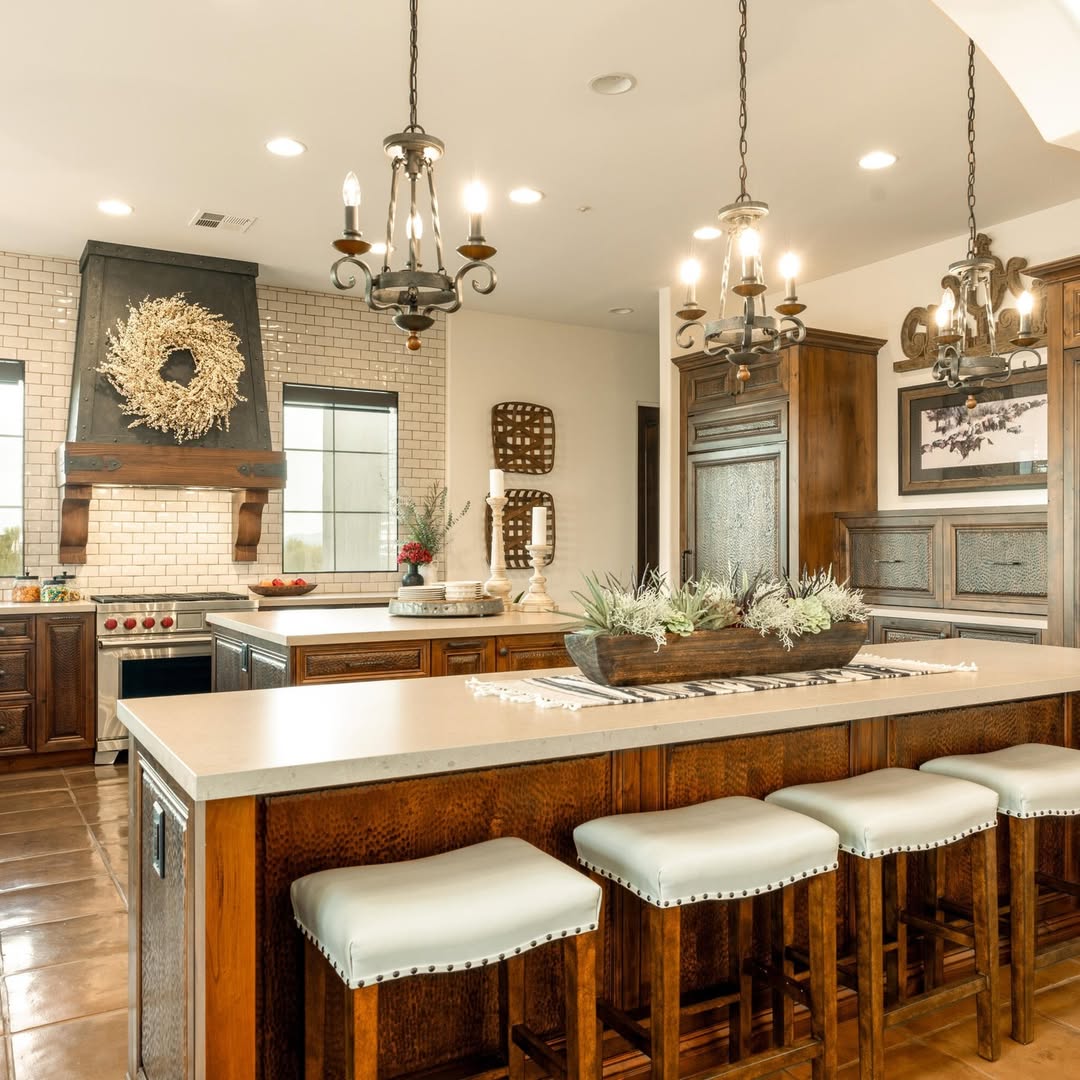 Spacious Mexican hacienda kitchen with wooden cabinets, large island with cushioned stools, modern appliances, tile flooring, and decorative lighting fixtures. A wreath hangs above the stove.