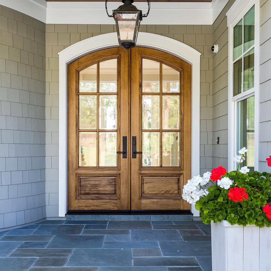 A stunning double door entryway with glass panels, framed by gray siding and a potted planter of red and white flowers, creates inspiring entryway ideas for any stone porch.