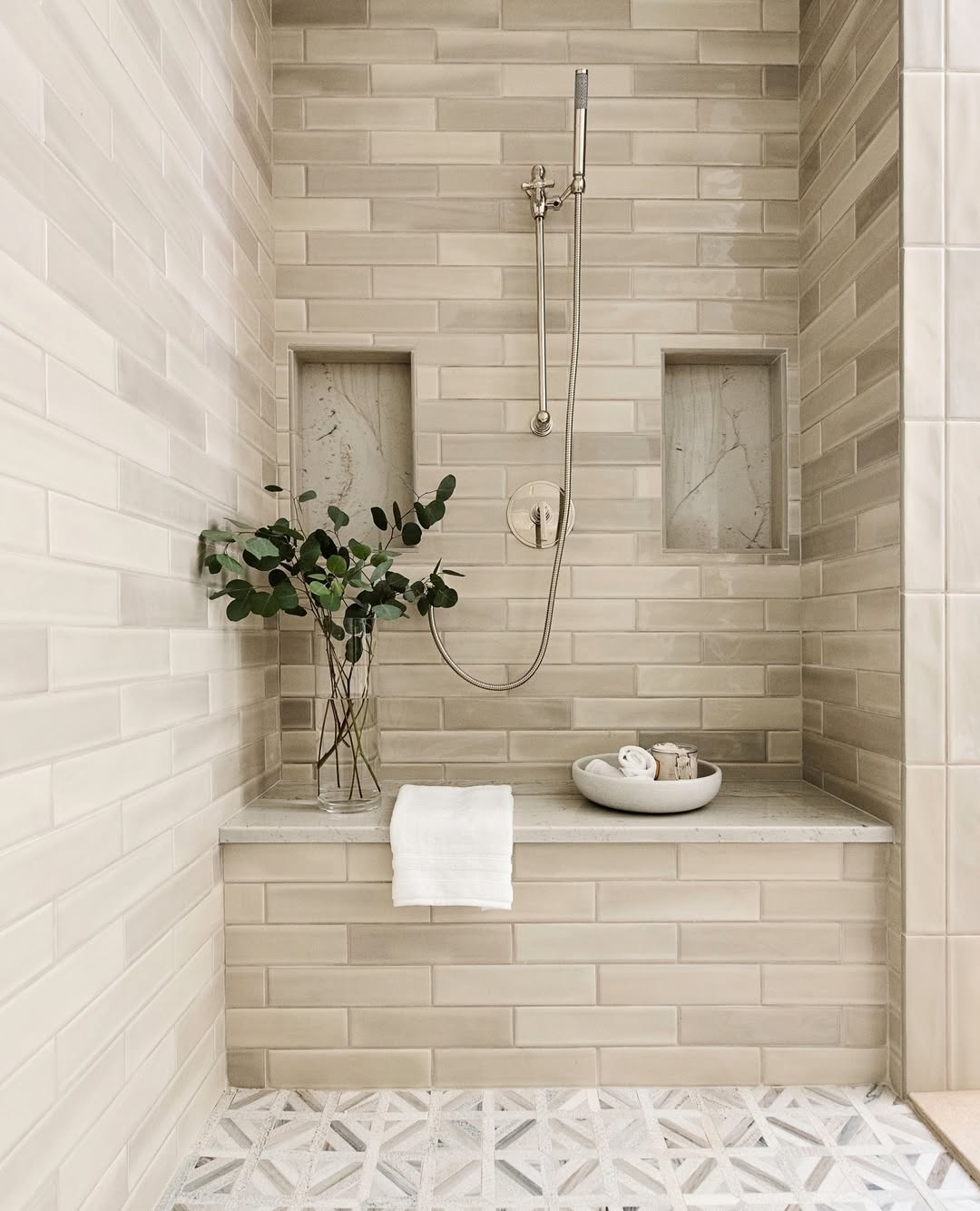 Modern shower with beige subway tiles showcases stylish bathroom tile ideas, featuring a built-in bench, two recessed shelves, a vase with greenery, a bowl with rolled towels, and a wall-mounted handheld showerhead for a true bathroom spa feel.