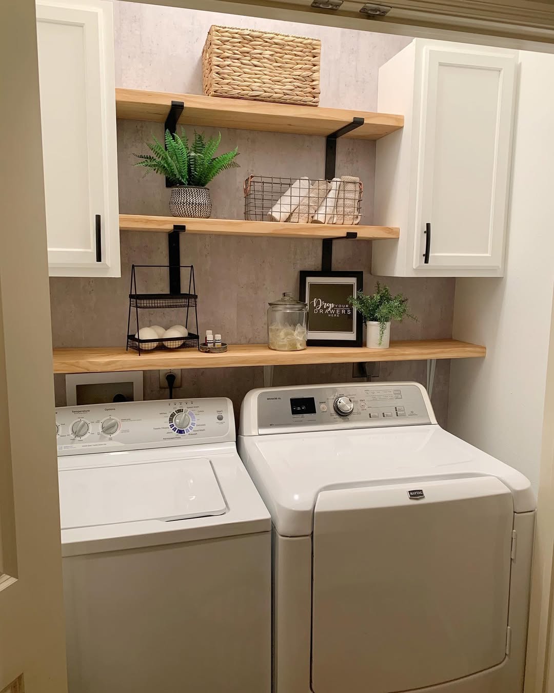A compact laundry room with a white washing machine and dryer, wooden shelves above holding baskets, boho decor, cleaning supplies, and potted plants—a perfect source for chic laundry room ideas.