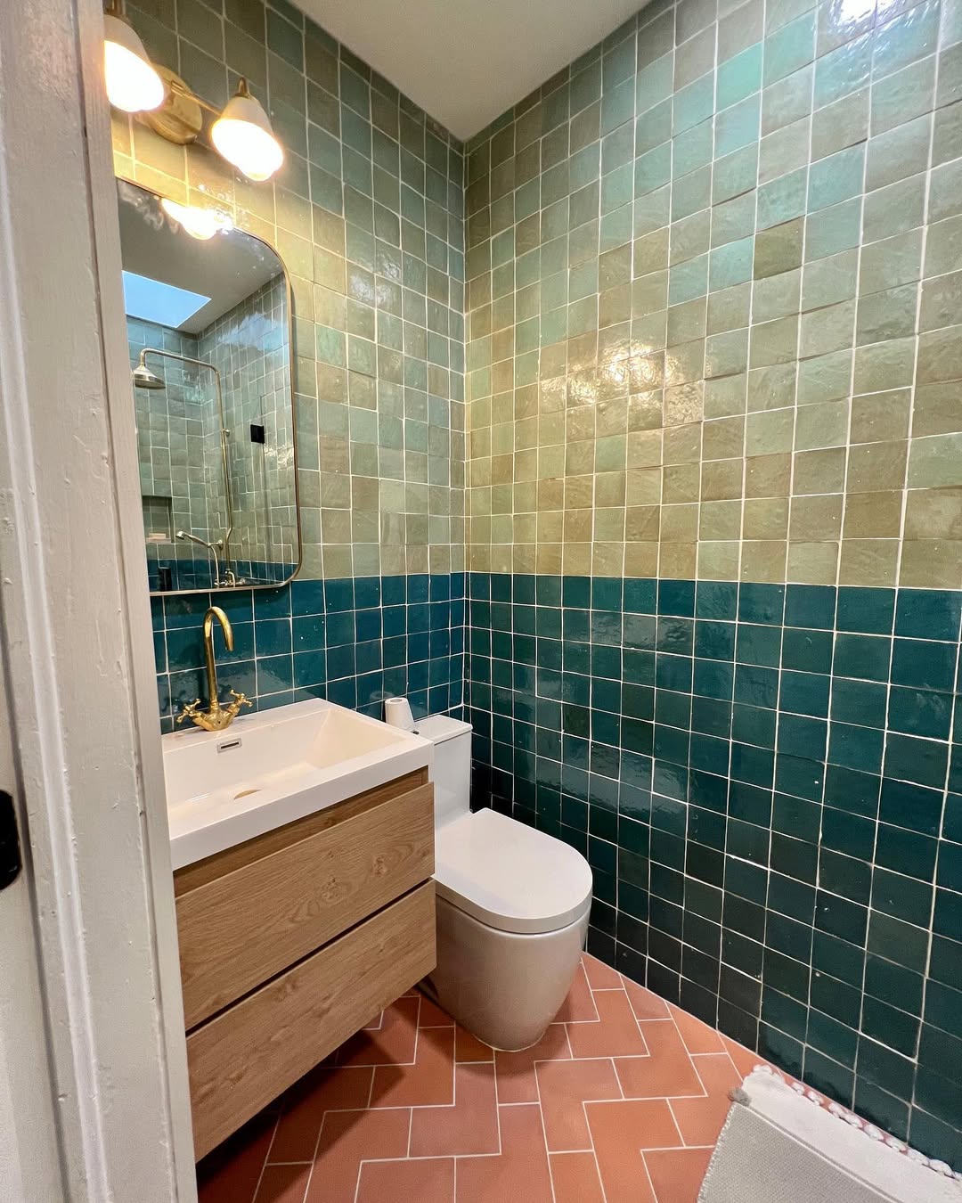 Small bathroom design with green and teal tiled walls, a wall-mounted mirror with lights, a wood vanity with sink, and a modern white toilet on a pink herringbone tile floor—perfect for spa-worthy bathroom inspiration.