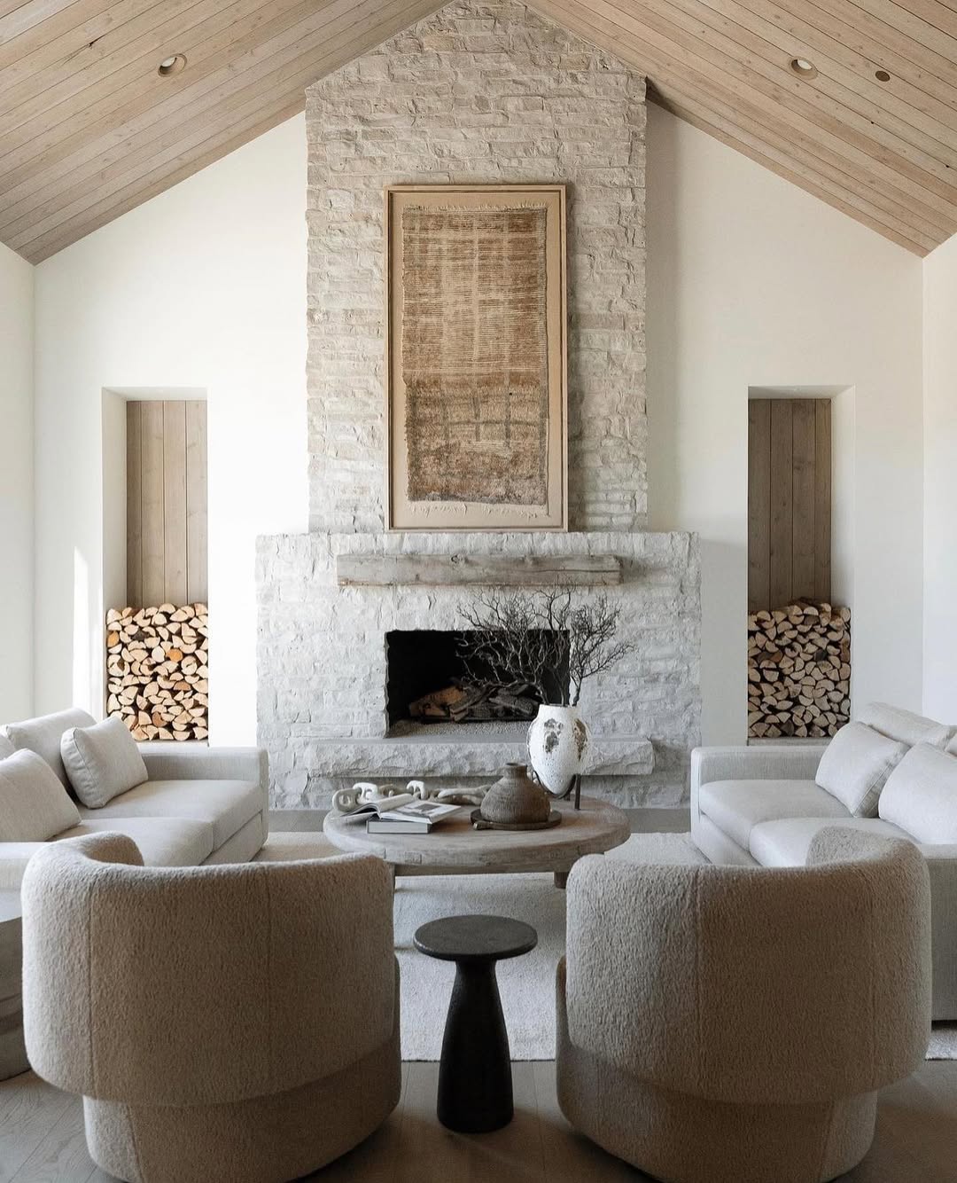 A modern organic living room with a stone fireplace, stacked firewood shelves, neutral-toned furniture, a round coffee table, and minimalist decor—perfect inspiration for expensive looking living rooms.