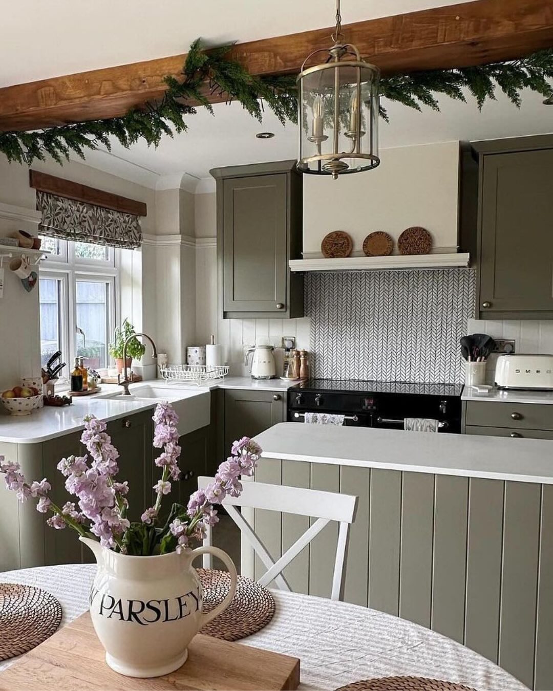 A cozy kitchen with sage green cabinets, white countertops, patterned backsplash, and a dining table with a