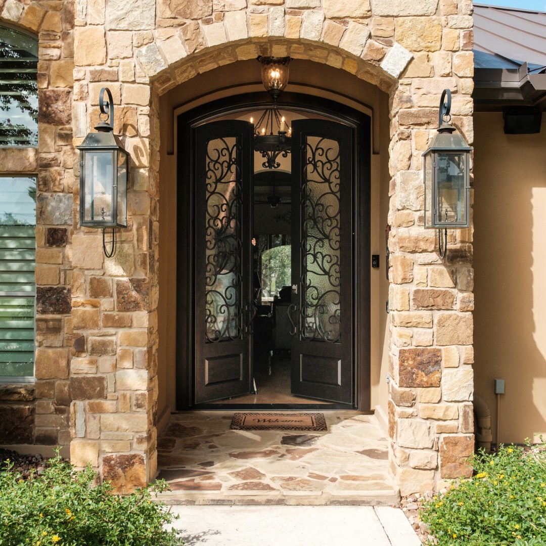 Stone entryway with two lantern-style wall lights, a stunning double door design featuring black wrought iron doors, and a welcome mat on a stone porch. A chandelier hangs above the entrance, offering timeless entryway ideas.