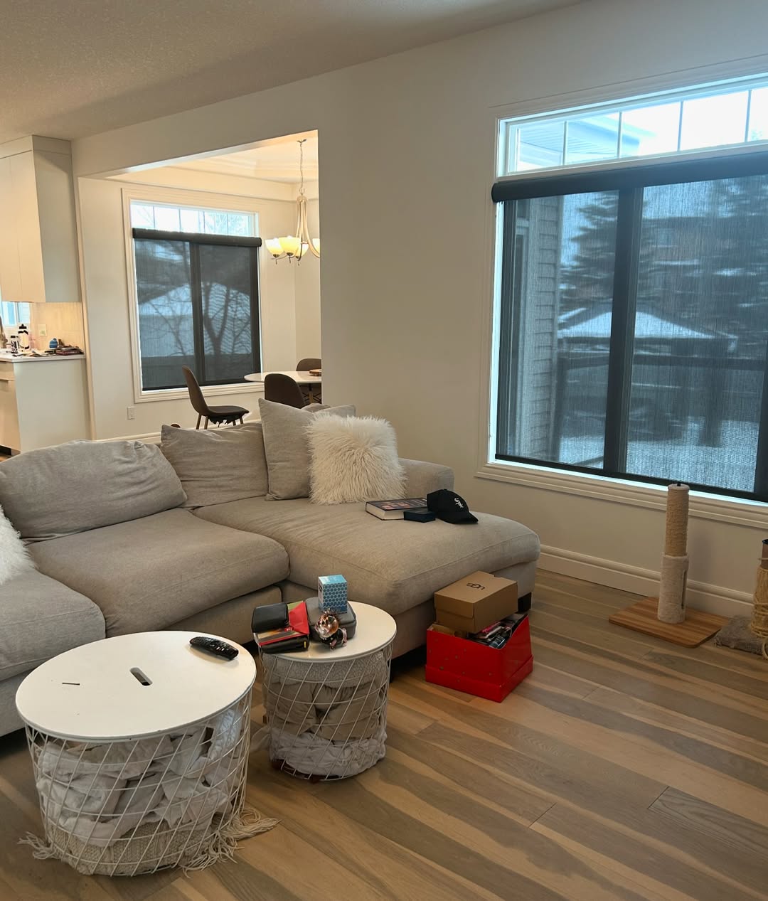 Modern living room ideas come to life with a grey sectional sofa, two round white tables, a cat scratching post, and large windows with black shades framing snowy views—perfect for an inviting organic modern living room ambiance.
