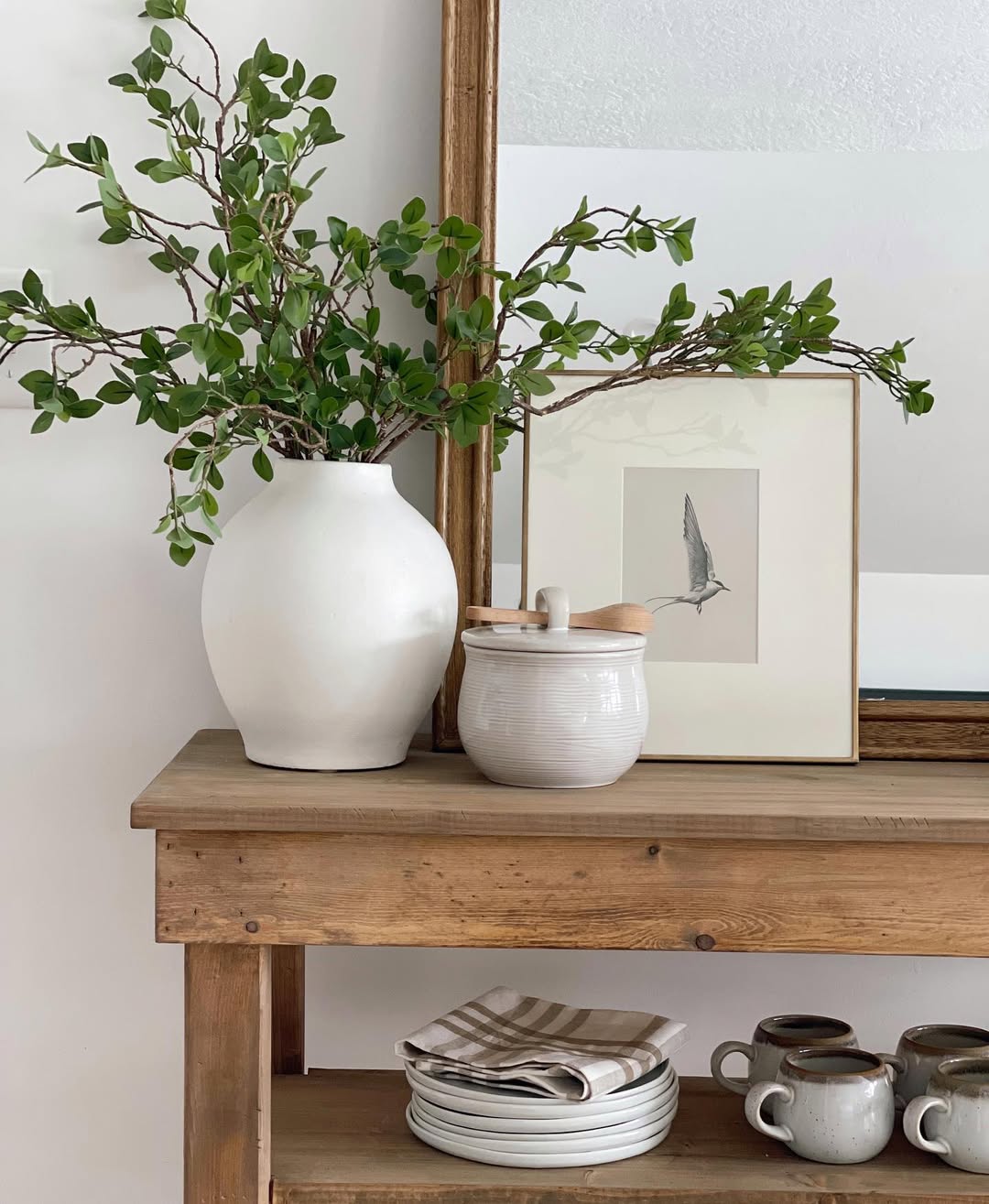 A wooden table with stacked plates, mugs, a striped cloth, creative vase decoration with green branches, a small lidded jar, and a framed bird sketch in front of a large mirror.