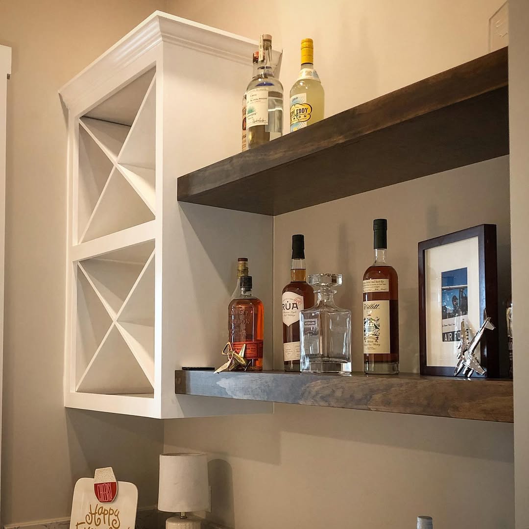 Two wooden shelves display assorted liquor bottles, a glass decanter, and a framed photo beside a stylish wine rack—perfect inspiration for those seeking practical wine rack ideas in a cozy home bar setting.