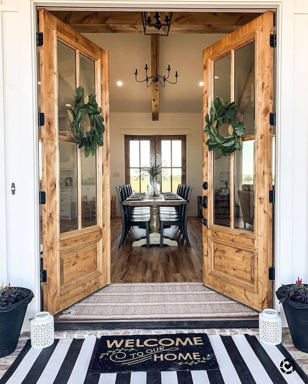 Double wooden doors with wreaths open to reveal a stylish double door entryway, leading to a dining room with a long table and striped chairs; a welcome mat reads