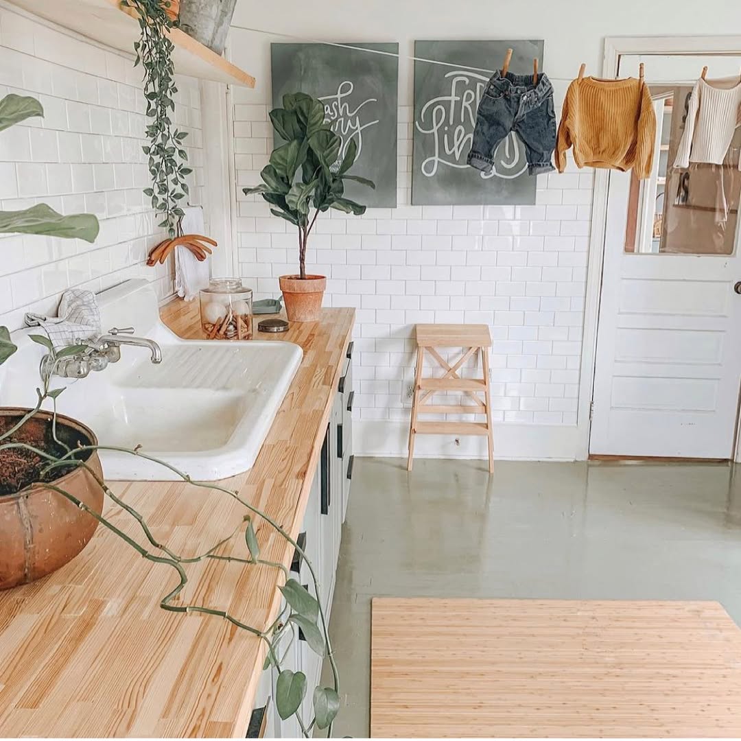 A Boho laundry room with a wooden countertop, white sink, potted plants, wall art, a stool, and clothes drying on a line above a white tiled wall and door showcases charming boho laundry decor.