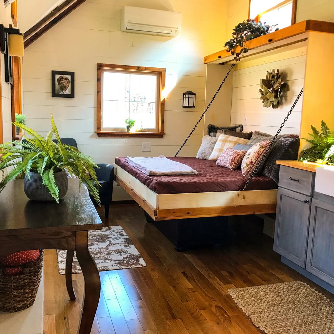 A cozy pet bedroom with a suspended bed, wooden floors, potted plants, a window, and a gray armchair next to a small table—perfect inspiration for delightful pet bedroom ideas.