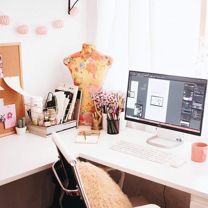 A tidy, well-lit home office workshop features a computer with design software open, a floral mannequin, stationery, books, a corkboard, a mug, and decorative lights on a white desk—perfect for inspiring new workshop ideas.