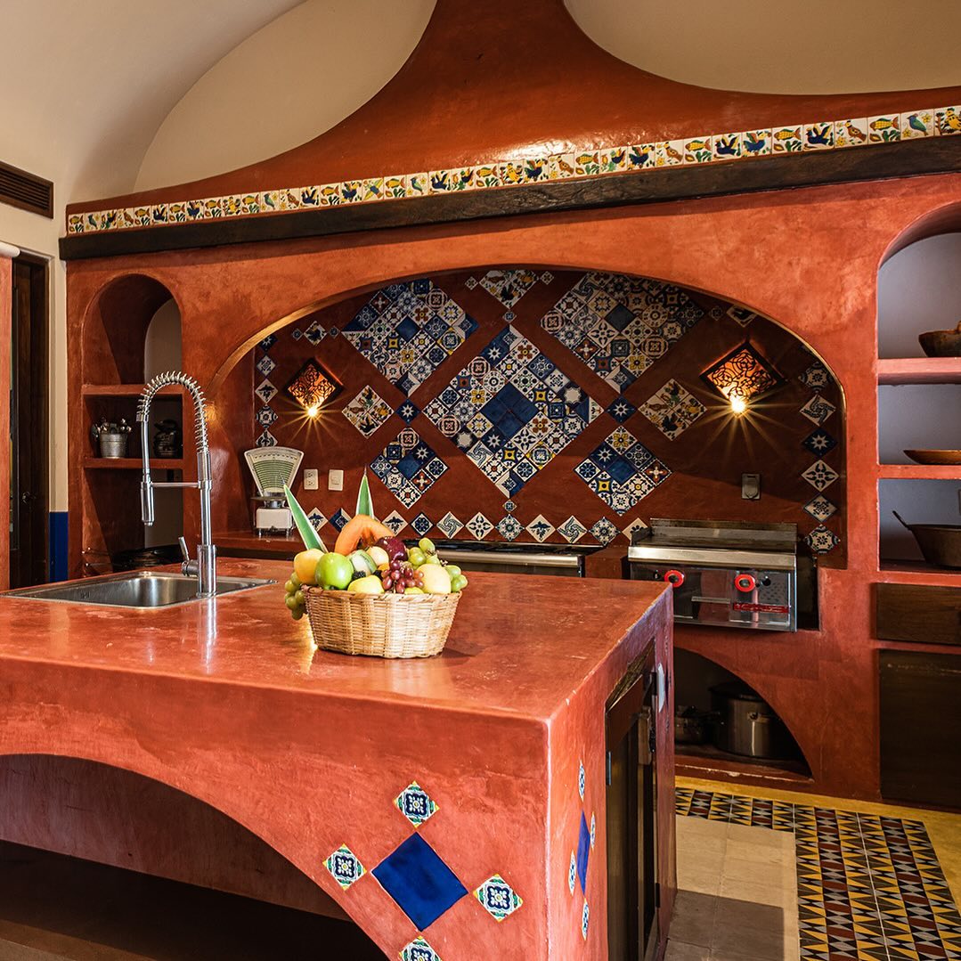 A vibrant Mexican hacienda kitchen with terracotta counters, blue and white patterned tiles, open shelving, a basket of fruit on the island, and a decorative hood above the stove area.