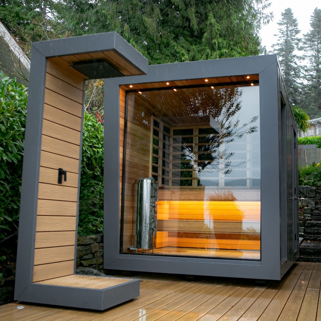 Modern outdoor sauna with large glass front, wooden interior, and gray exterior frame, situated on a wooden deck surrounded by greenery—an inspiring choice for anyone seeking unique outdoor sauna ideas.