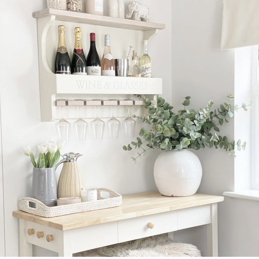 A home bar setup featuring stylish wine storage shelves holding wine and liquor bottles, hanging wine glasses, a tray with cups, and a large vase of greenery on a white wooden table.