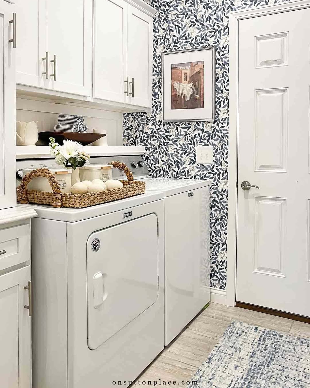 A laundry room with a white washer and dryer, stylish Laundry Room Cabinet Ideas, a wicker tray with decor, floral wallpaper, a framed picture, and a patterned rug.