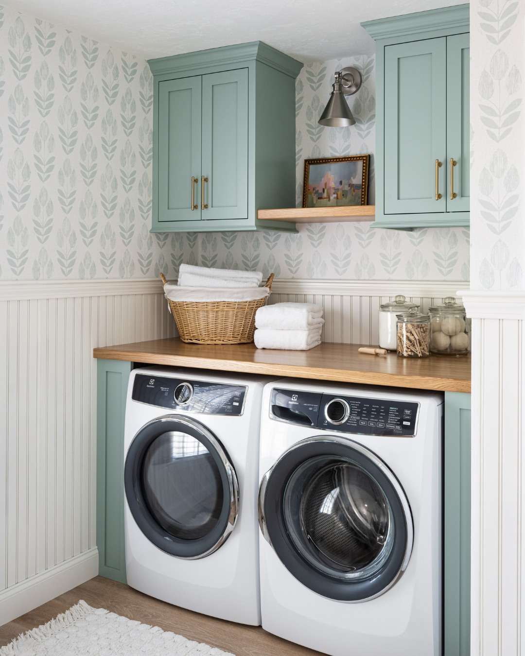 A laundry room with stylish Laundry Room Cabinets, a front-loading washer and dryer, green cabinetry, a wooden countertop, a basket with towels, and a decorative shelf above.